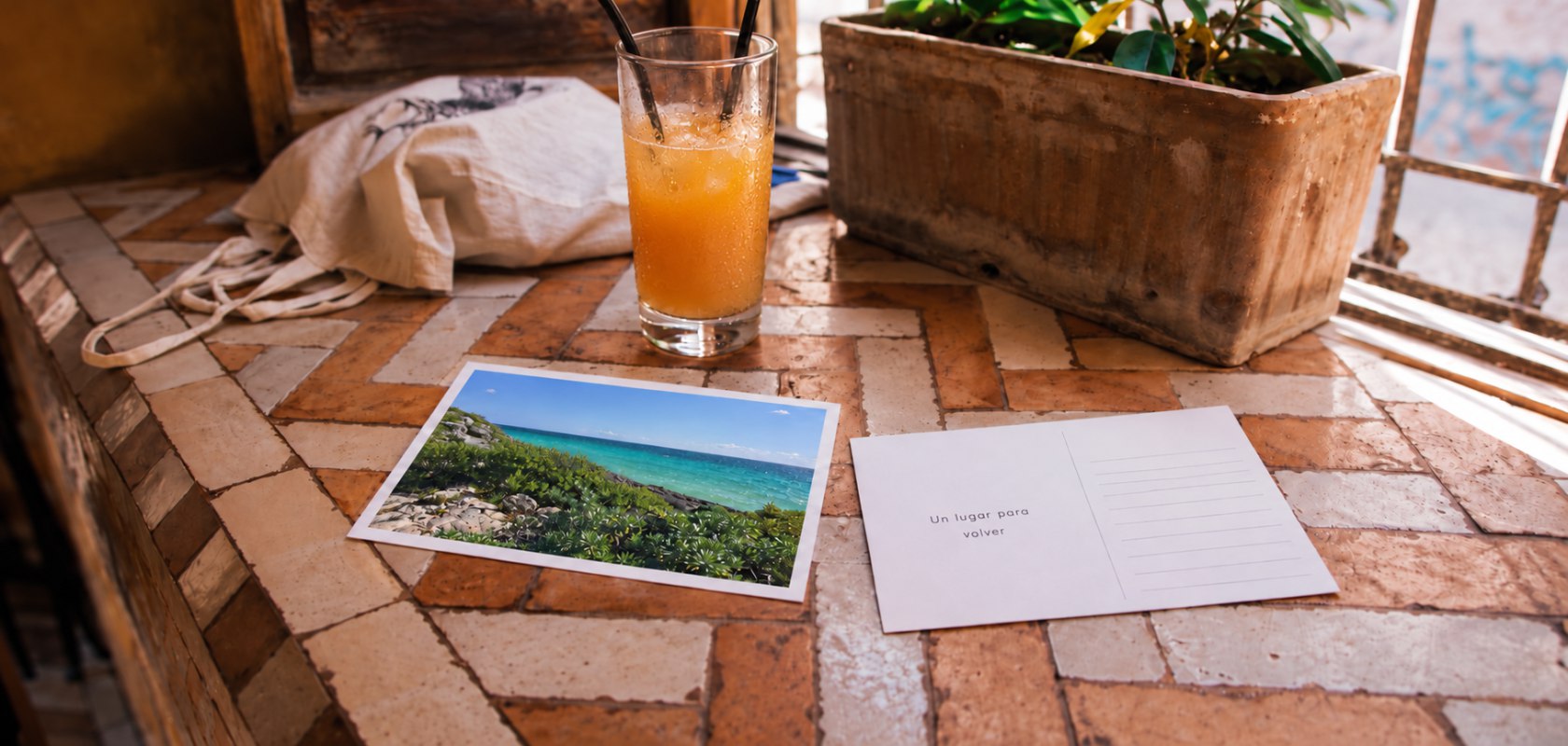 Frente con un paisaje y dorso con dedicatoria de dos postales personalizadas 10 por 15 sobre una mesa de baldosas, junto a un vaso de jugo.